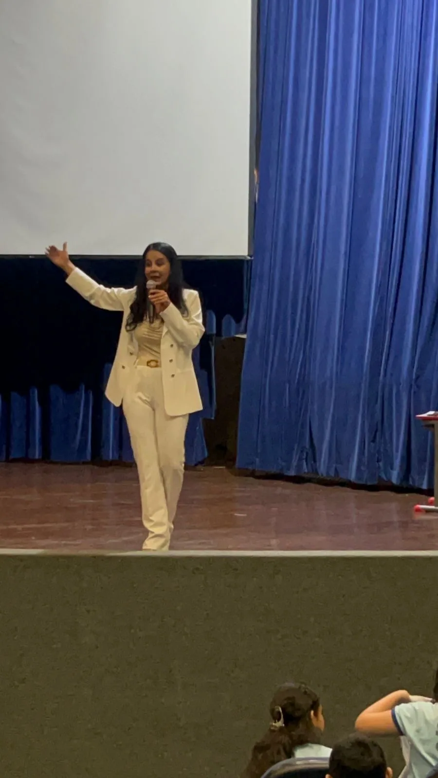 Dra. Genaina Morais falando ao microfone durante uma palestra, em palco com cortina azul